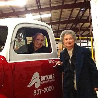 Older couple with vintage red-and-white Butcher Air Conditioning truck in warehouse, woman pointing at logo