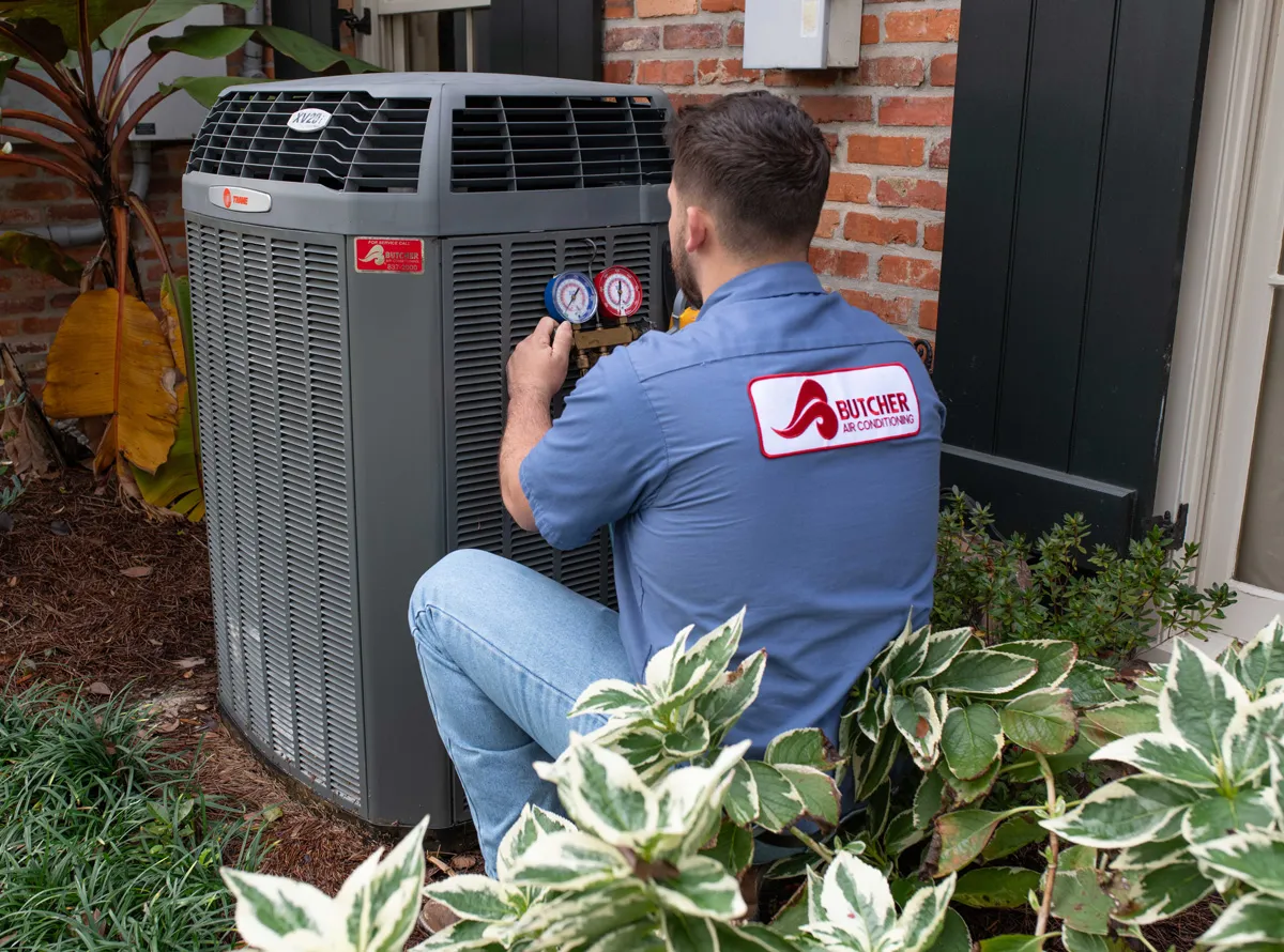 HVAC technician inspecting outdoor air conditioner unit with gauges, wearing Butcher Air Conditioning uniform