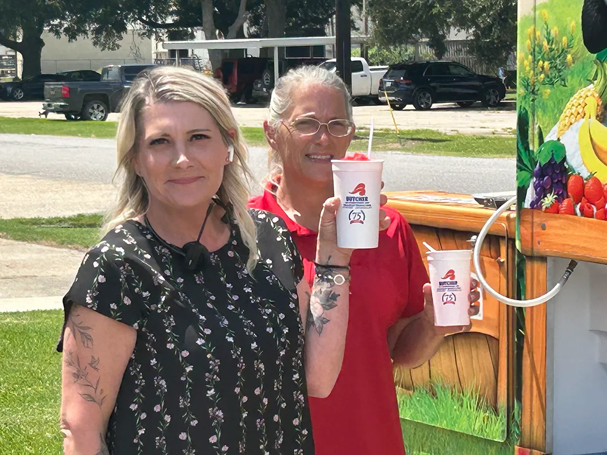 Two women smiling and holding frozen drink cups in front of colorful shaved-ice food truck on a sunny day
