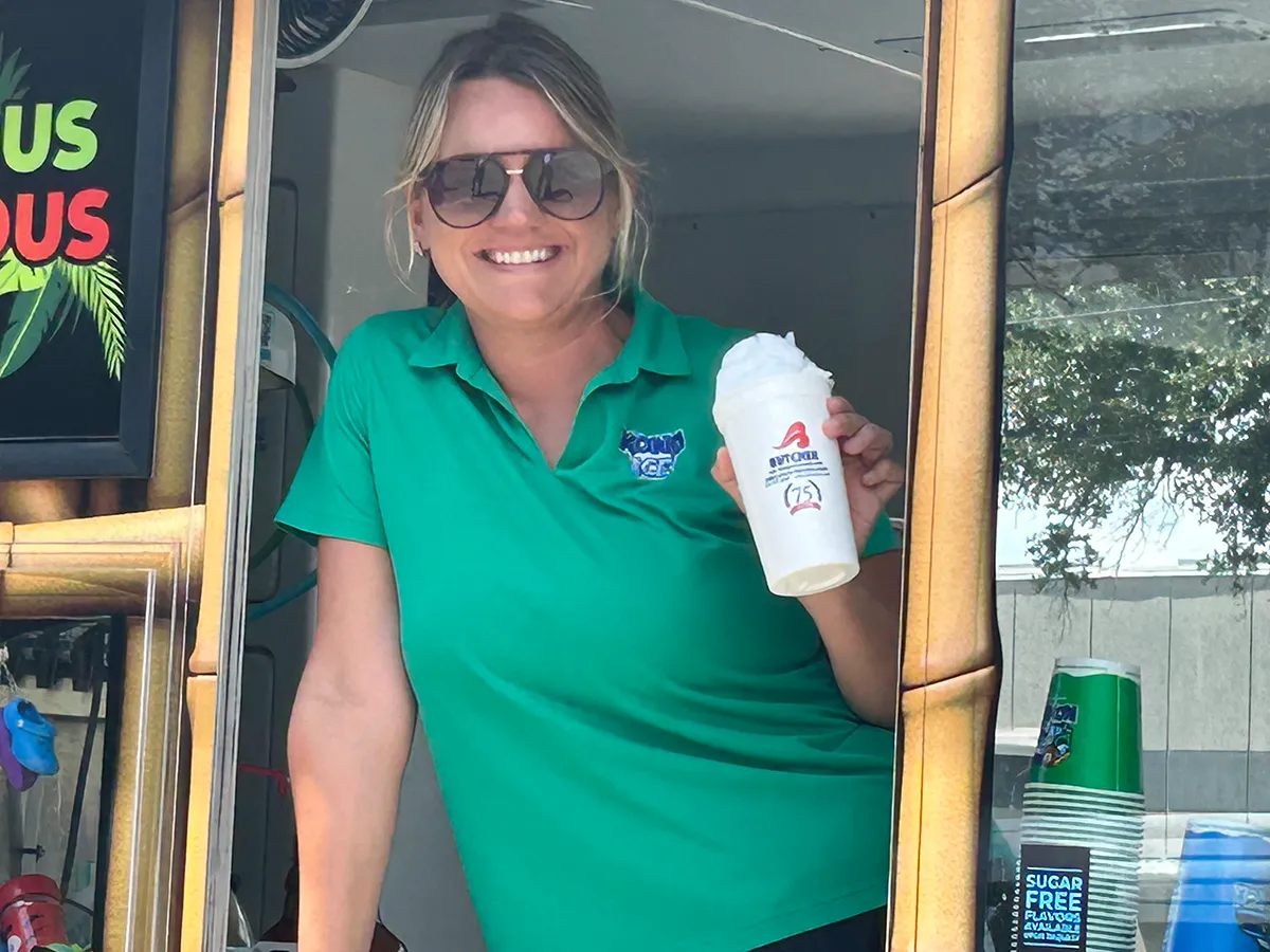 Smiling woman in green polo serving a frozen drink from a food-truck window, holding a white cup