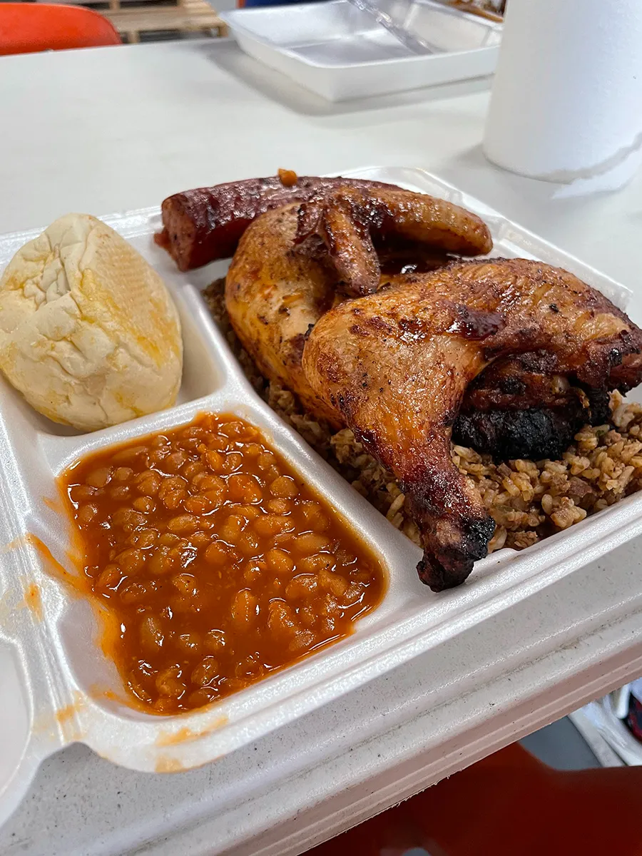 BBQ chicken quarter with smoked sausage on seasoned rice, served with baked beans and a bread roll in foam container