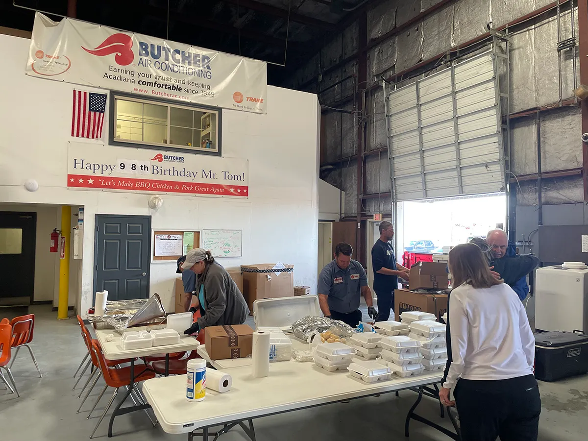 Workers packing takeaway meals on folding tables in a Butcher Air Conditioning warehouse beneath birthday banner