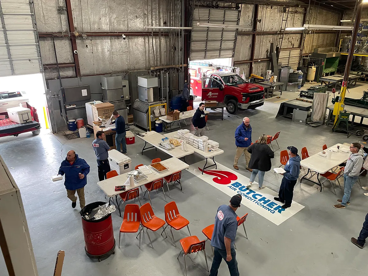 HVAC technicians eating lunch in industrial shop with Butcher Air Conditioning logo, tables, food trays and red service truck