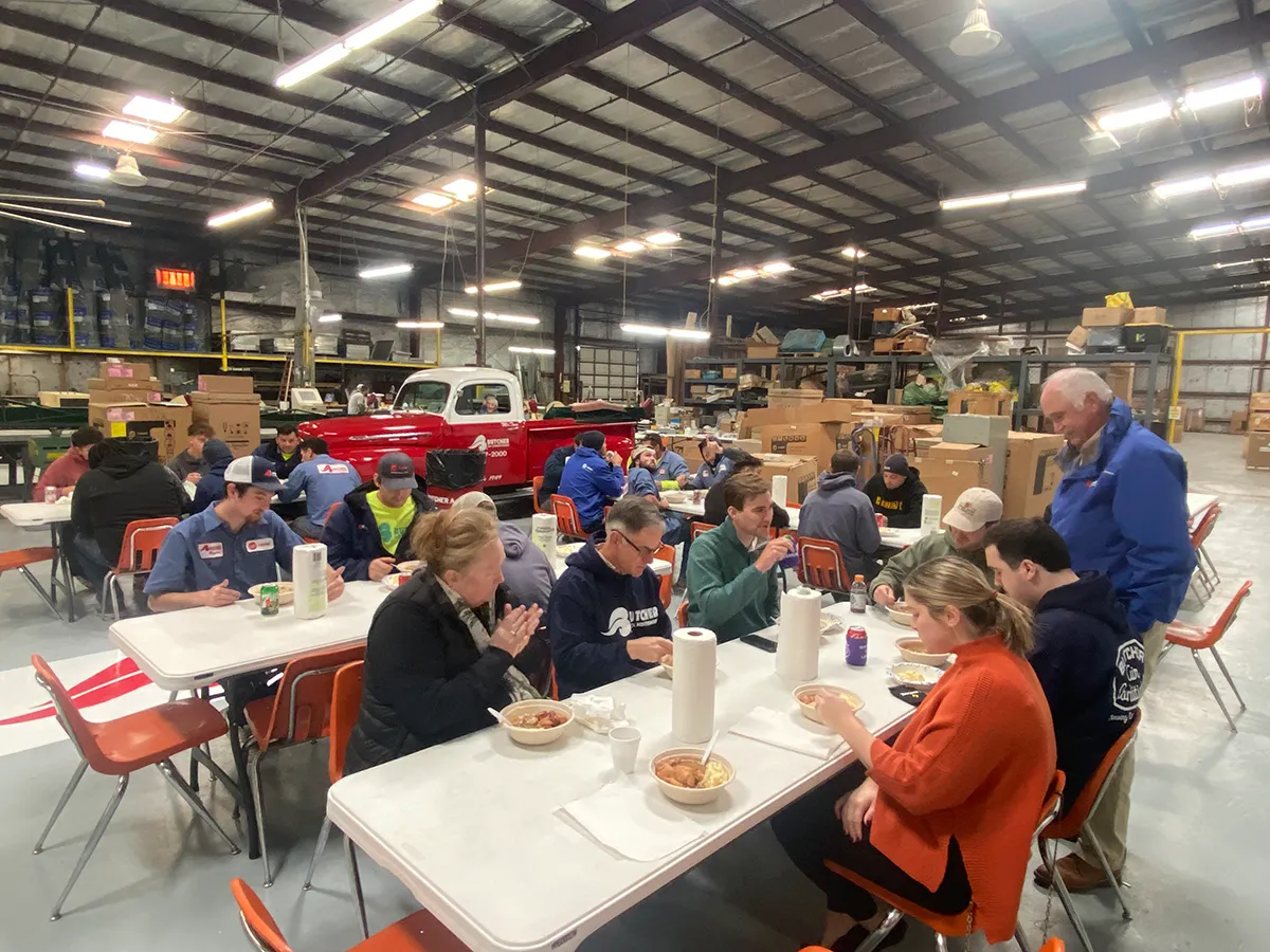 Workers eating lunch at long tables inside a warehouse with stacked boxes and a red vintage pickup truck in background