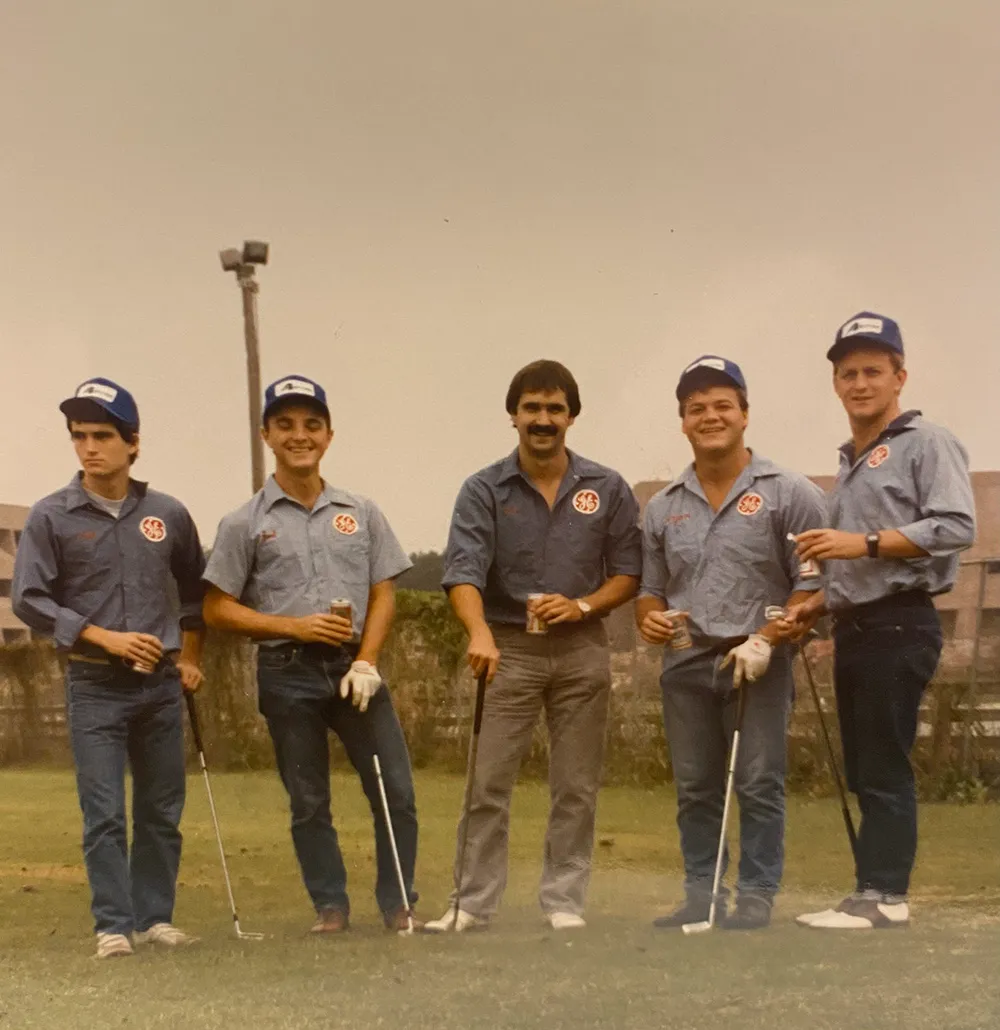Five men in matching blue shirts and caps holding golf clubs and beers on a golf course