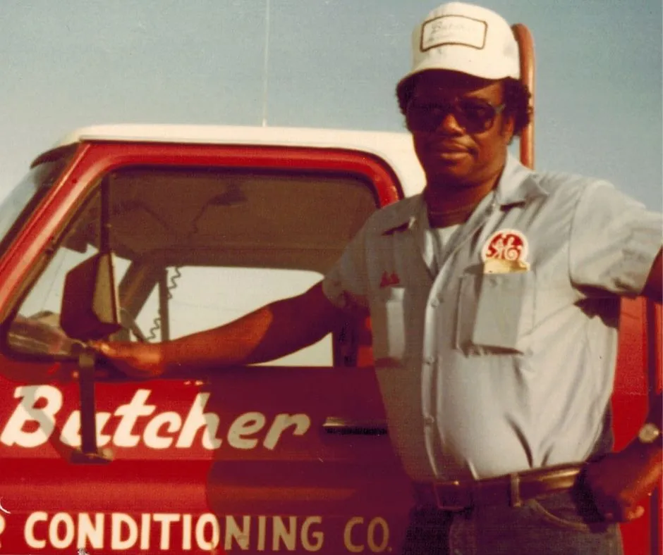 Man in work uniform and cap leaning on red 'Butcher Air Conditioning Co.' truck, vintage HVAC technician.
