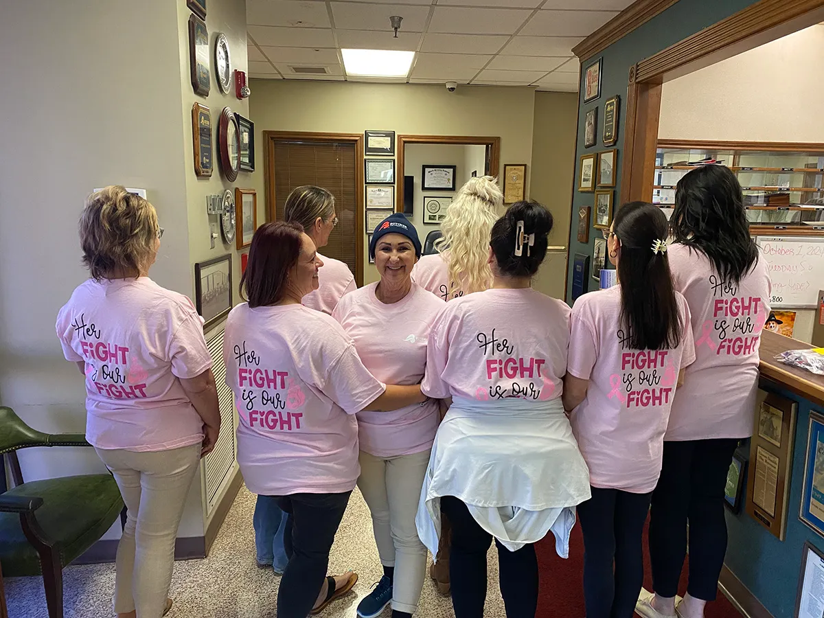 Group of women wearing pink 'Her Fight is Our Fight' shirts supporting breast cancer awareness in an office.