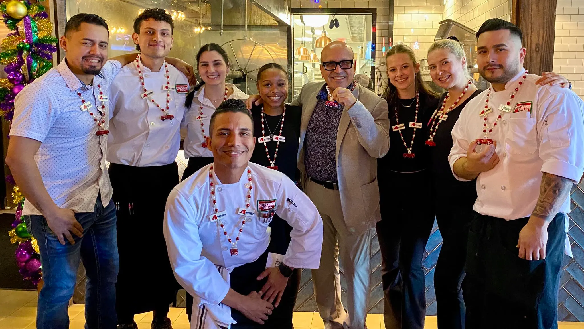 Smiling restaurant staff and owner posing in kitchen with holiday beaded necklaces, chefs wearing Superior Grill jackets