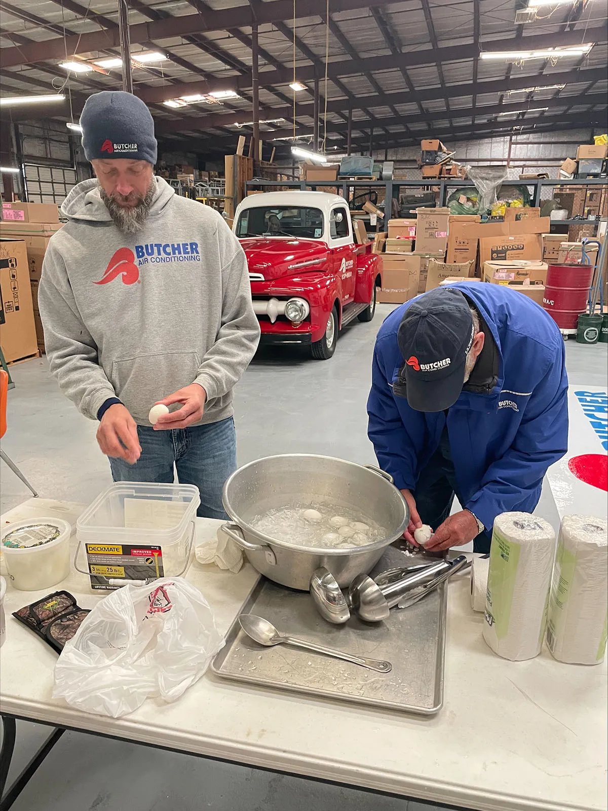 Two workers in Butcher Air Conditioning gear peeling hard-boiled eggs at a table in a warehouse with a vintage red pickup
