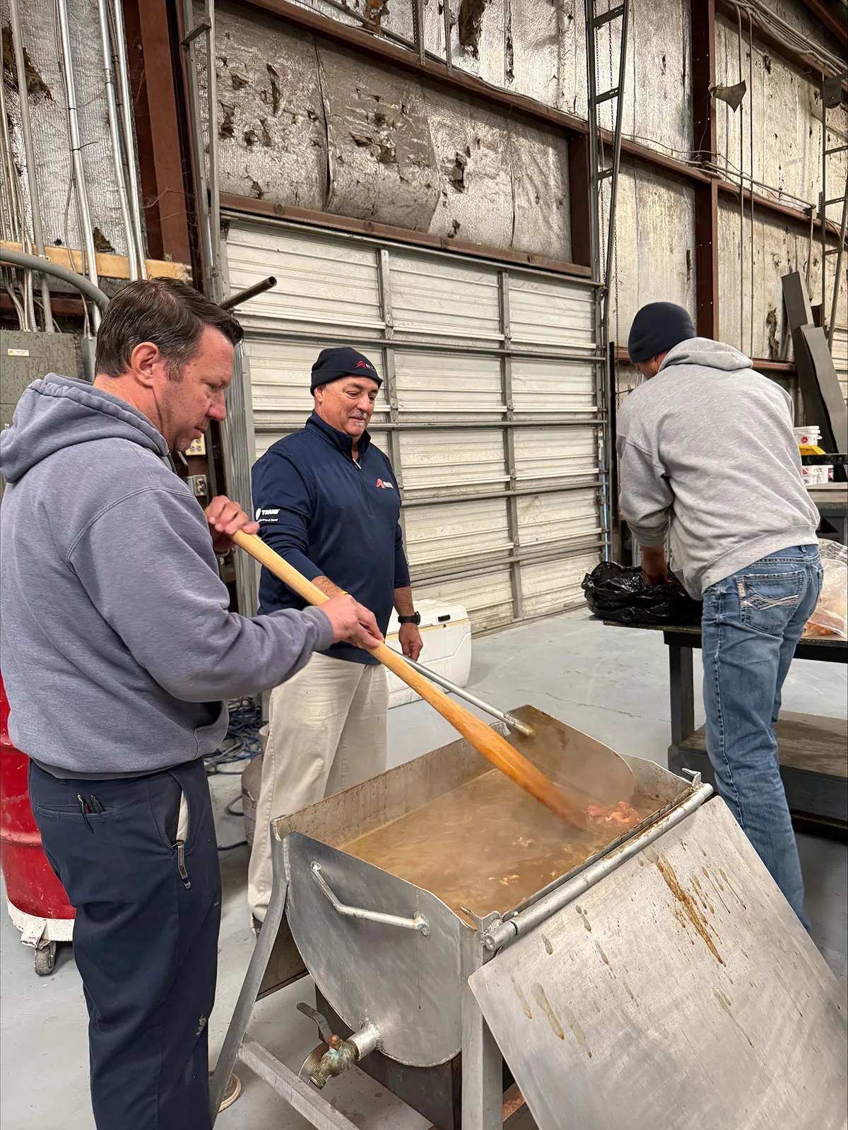 Three men cooking a large batch of stew in a metal kettle inside an industrial garage with overhead door