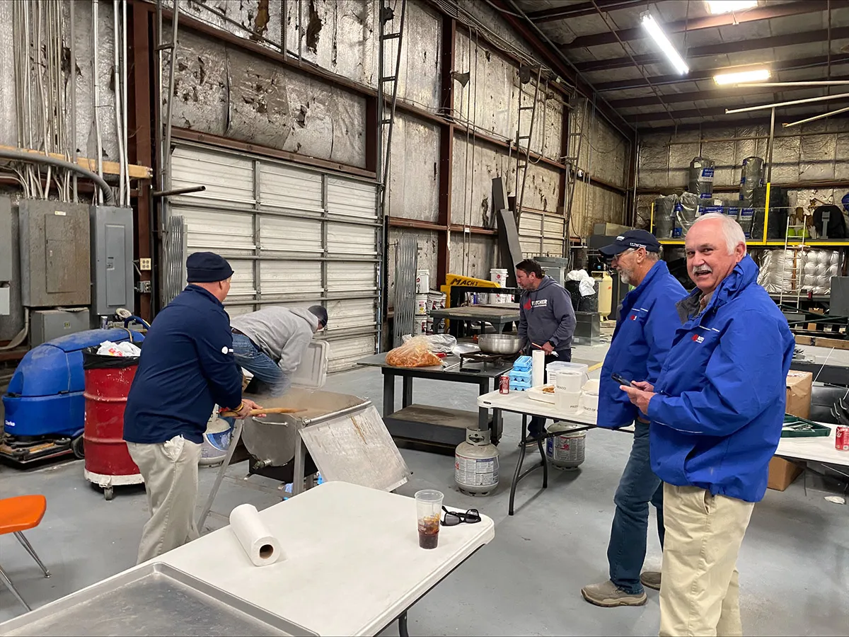 Men in blue jackets cooking and serving food on portable burners and folding tables inside an industrial warehouse