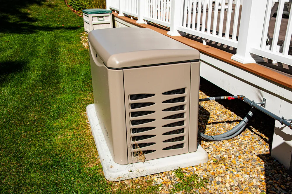 Residential standby generator on concrete pad beside house with utility connections, gravel bed and white porch railing