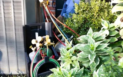 HVAC technician servicing outdoor air conditioner unit with manifold gauges and refrigerant hoses beside a brick house.