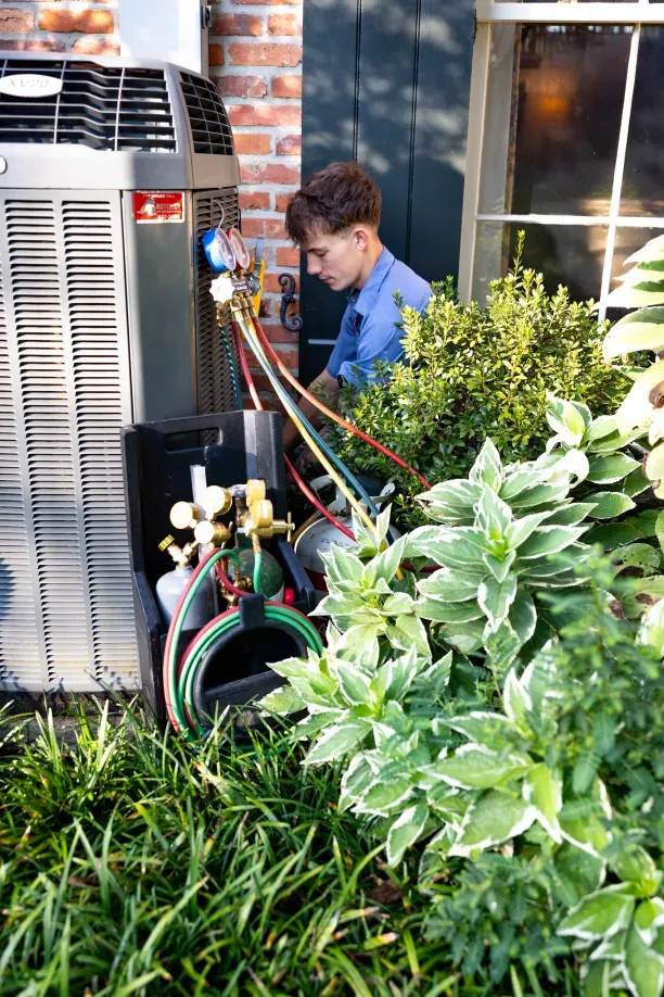 HVAC technician servicing outdoor air conditioner unit with manifold gauges and refrigerant hoses beside a brick house.