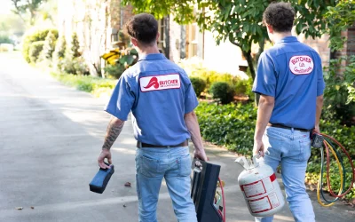 Two HVAC technicians in Butcher Air Conditioning uniforms carrying refrigerant tank and tool cases on a residential driveway