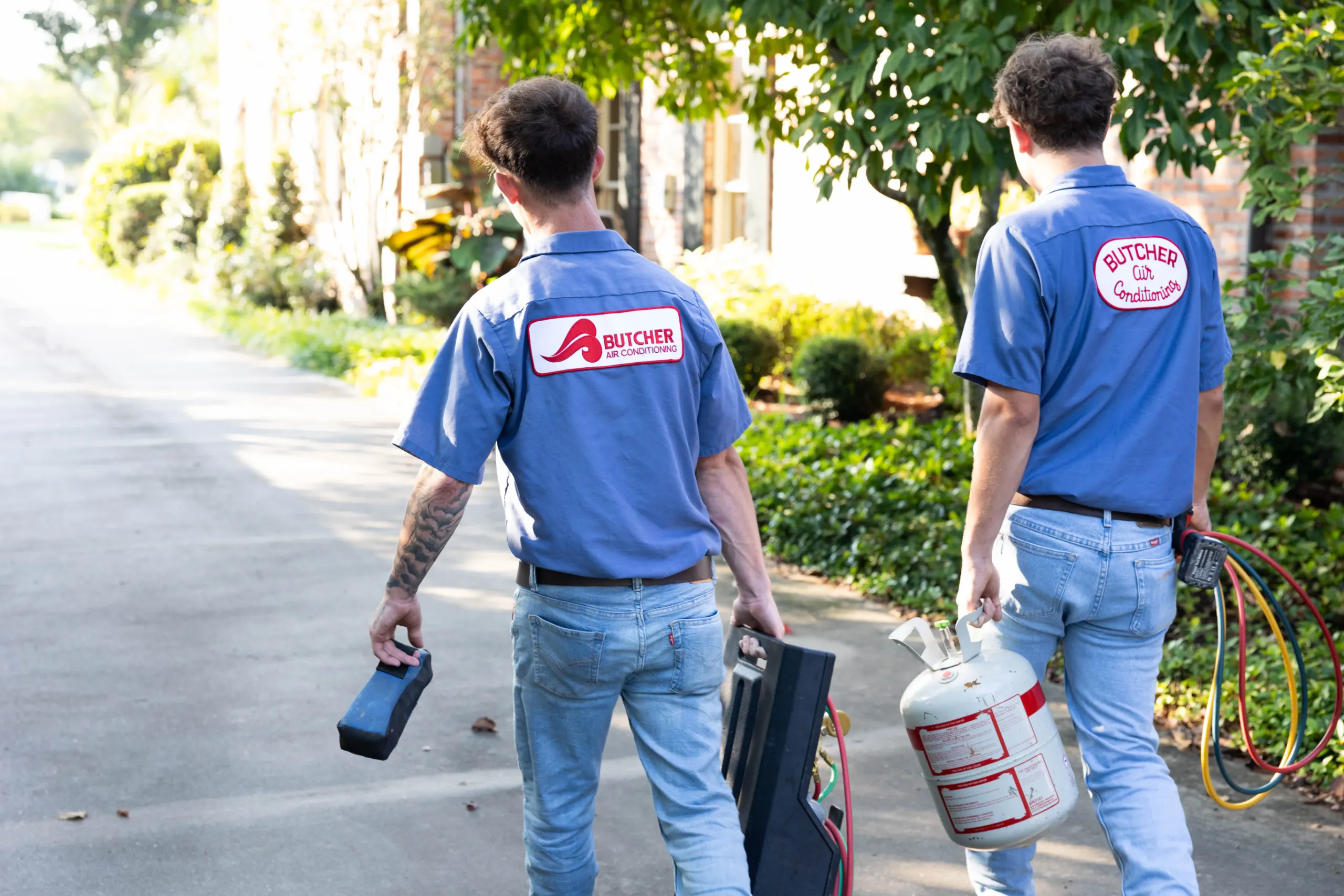Two HVAC technicians in Butcher Air Conditioning uniforms carrying refrigerant tank and tool cases on a residential driveway