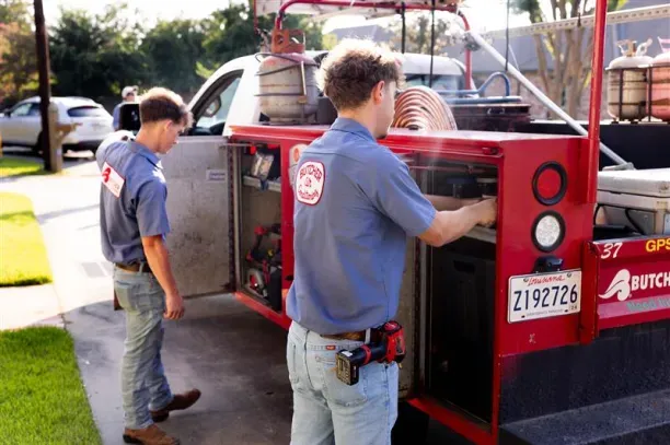 Two technicians in blue shirts accessing equipment compartments on a red service truck in a suburban driveway