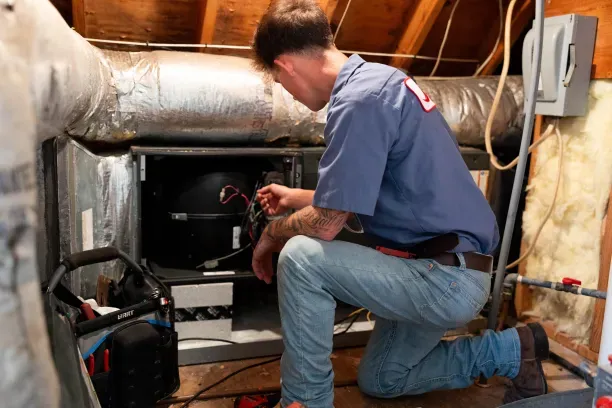 A man kneeling in an attic HVAC unit repairing an air handler, inspecting wiring with toolbox nearby
