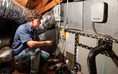 HVAC technician kneeling in attic testing air handler with multimeter near ductwork and control panels
