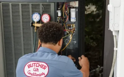 HVAC technician in Butcher Air Conditioners shirt servicing outdoor AC unit with pressure gauges and wiring.