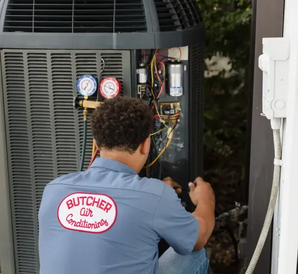 HVAC technician in Butcher Air Conditioners shirt servicing outdoor AC unit with pressure gauges and wiring.