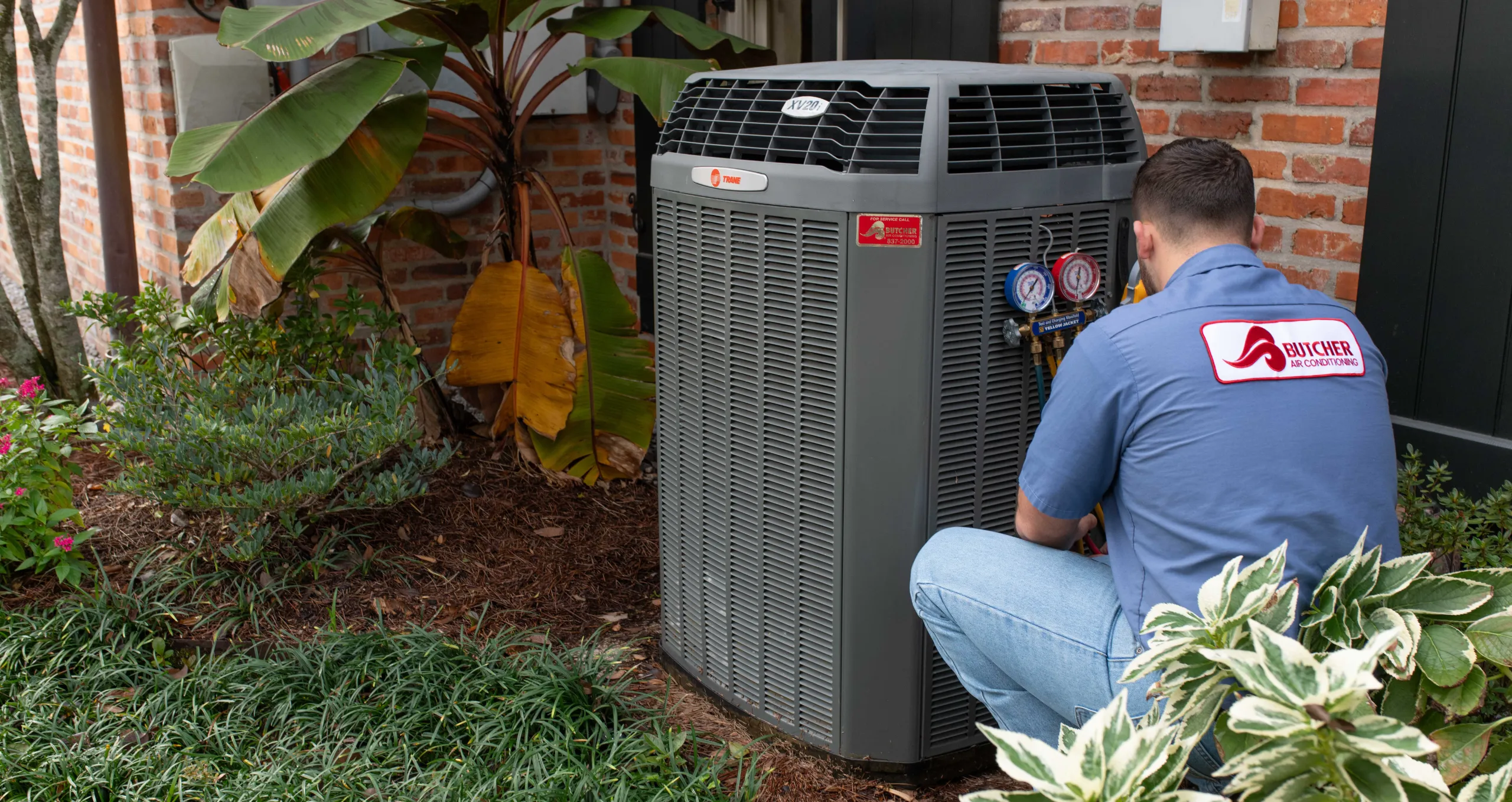 A man HVAC technician servicing outdoor air conditioning condenser unit with pressure gauges by a brick house