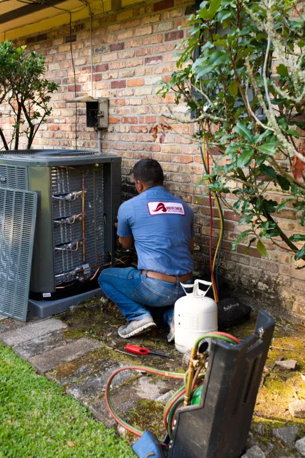A man kneeling by outdoor air conditioner unit repairing HVAC system with refrigerant tank and manifold gauges.