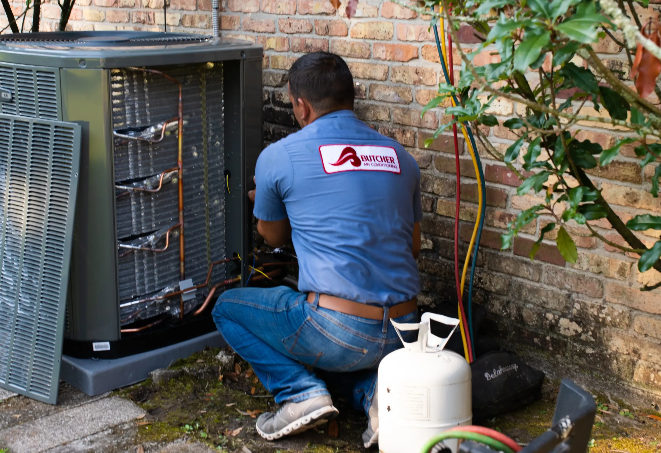 HVAC technician repairing outdoor air conditioner next to brick wall with refrigerant tank and service hoses