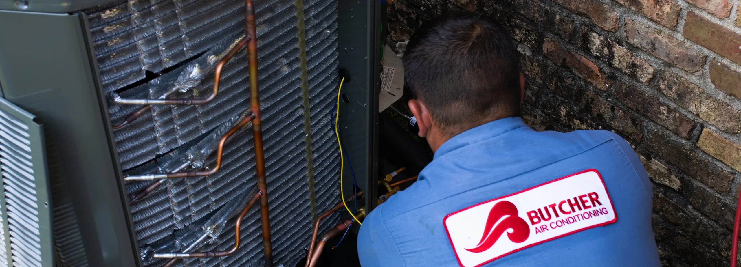 HVAC technician repairing outdoor air conditioner condenser coils and copper tubing beside brick wall