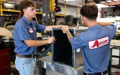 Two HVAC technicians in blue Butcher Air Conditioning shirts assembling metal ductwork on a workbench in a fabrication shop