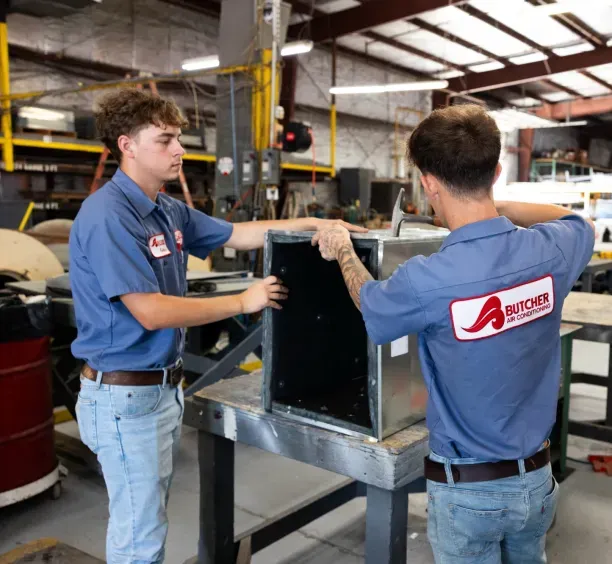 Two HVAC technicians in blue Butcher Air Conditioning shirts assembling metal ductwork on a workbench in a fabrication shop