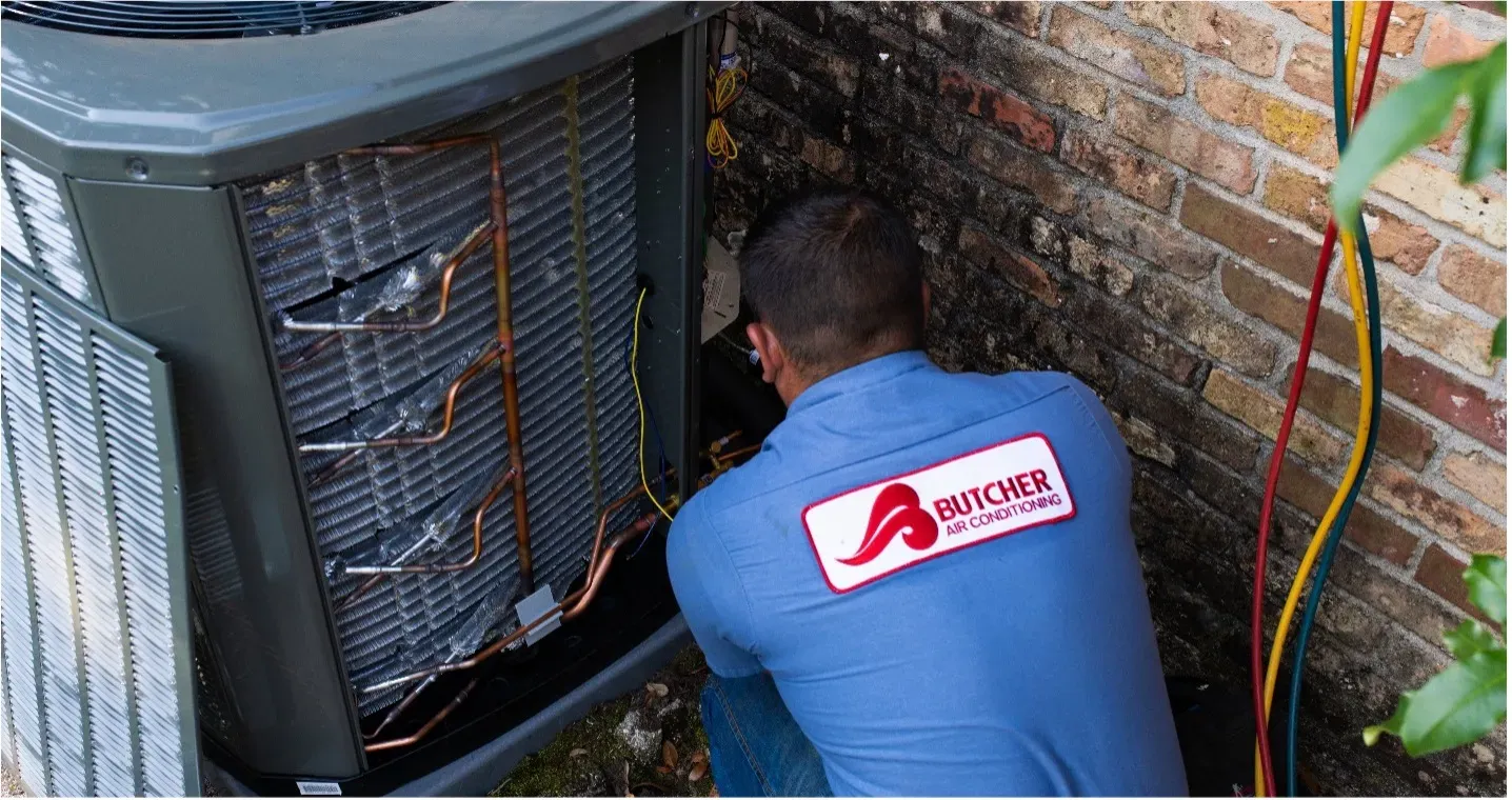 HVAC technician repairing outdoor air conditioning condenser with exposed copper coils and refrigerant hoses by brick wall