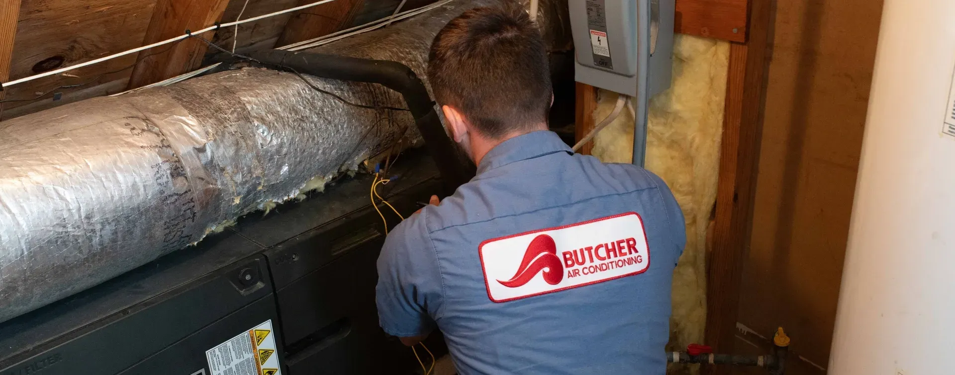 HVAC technician wearing Butcher Air Conditioning shirt repairs insulation-wrapped duct and air handler in attic